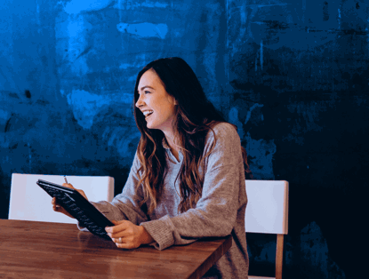 Vrouw in grijze trui zit aan een houten tafel en kijkt naar een tablet, met een blauwe muur op de achtergrond.