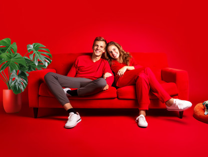 Twee mensen in rode kleding zitten samen op een felrood bankstel, met een groene plant op de achtergrond.