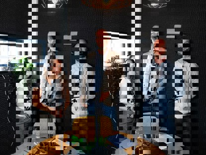 Drie mensen met een microfoon in een opnamestudio voeren een gesprek. Op de tafel staat een glas water en een notitieblad.