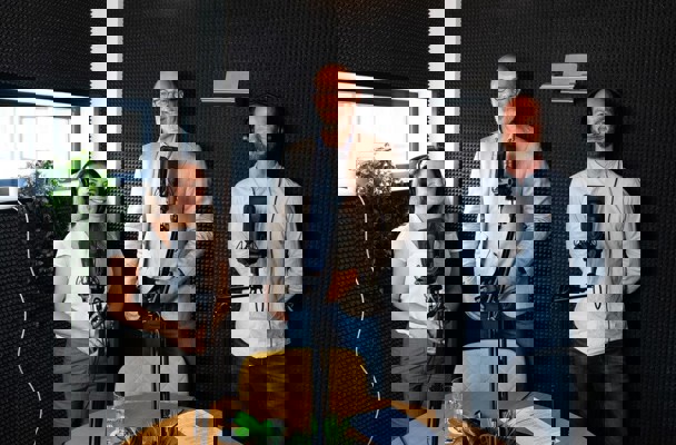 Drie mensen met een microfoon in een opnamestudio voeren een gesprek. Op de tafel staat een glas water en een notitieblad.