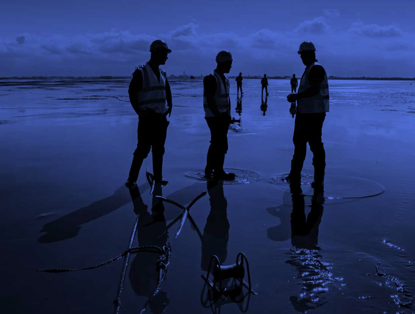Vijf personen in veiligheidsvesten staan op nat zand met reflecties en bespreken een samenwerkingsproject.
