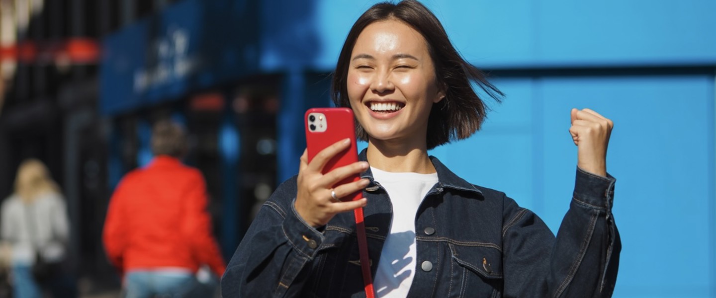 Persoon in denimjack juicht terwijl hij een smartphone vasthoudt, met een blauwe muur op de achtergrond.