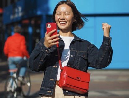 Persoon in denimjack juicht terwijl hij een smartphone vasthoudt, met een blauwe muur op de achtergrond.