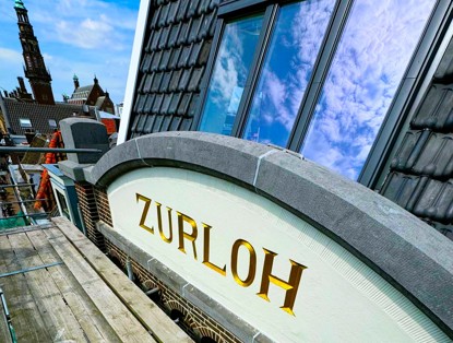 Sign with the text ZURLOH in gold letters, positioned above a window on a building, against a backdrop of bright blue sky.