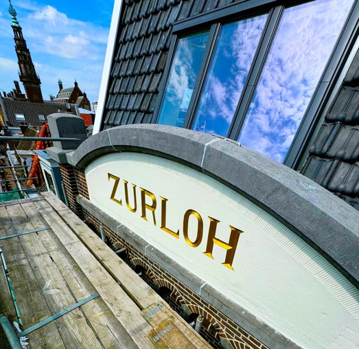 Bord met de tekst ZURLOH in gouden letters, boven een raam op een gebouw, tegen een heldere blauwe lucht.