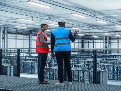 Two men in safety gear, one wearing a red vest and the other a blue vest, are observing a data center filled with servers.