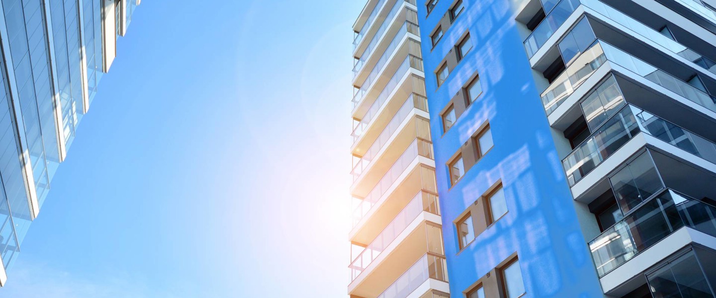 Tall building with glass balconies and a blue facade, against a clear blue sky with sunlight.