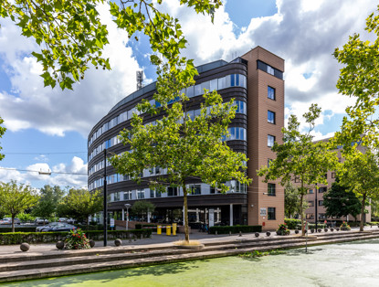 Modern office building with large windows and a round design, surrounded by lush trees and a water feature.