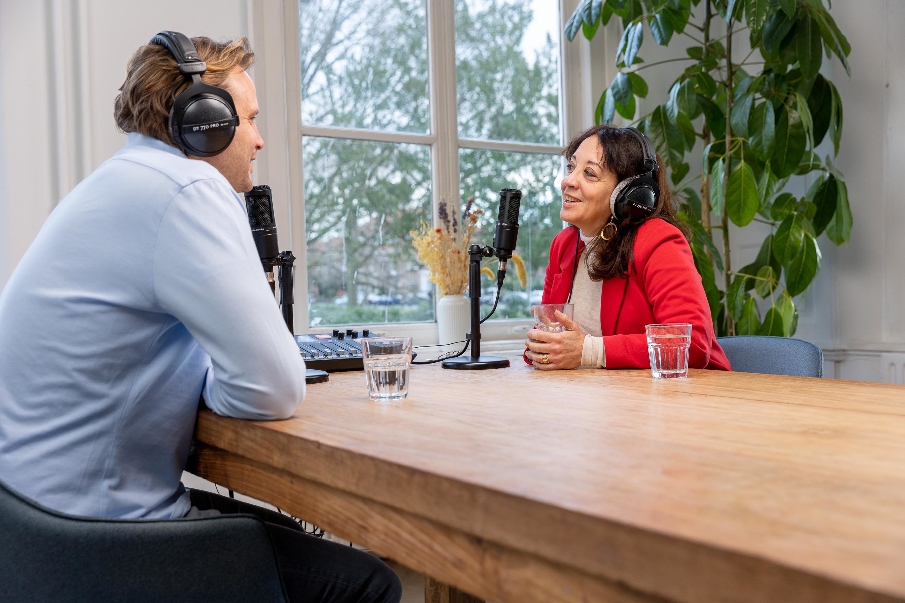 Twee mensen converseren aan een houten tafel, beiden met een microfoon en hoofdtelefoons, in een lichte ruimte omringd door planten.