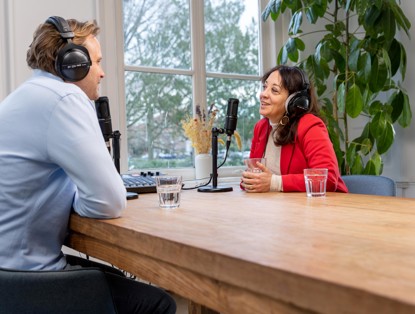 Twee mensen converseren aan een houten tafel, beiden met een microfoon en hoofdtelefoons, in een lichte ruimte omringd door planten.