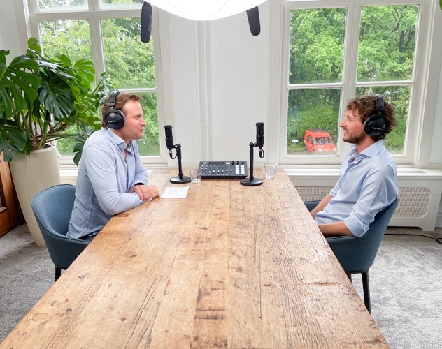 Twee mannen zitten tegenover elkaar aan een houten tafel met microfoons. Op de achtergrond zijn groene bomen te zien.