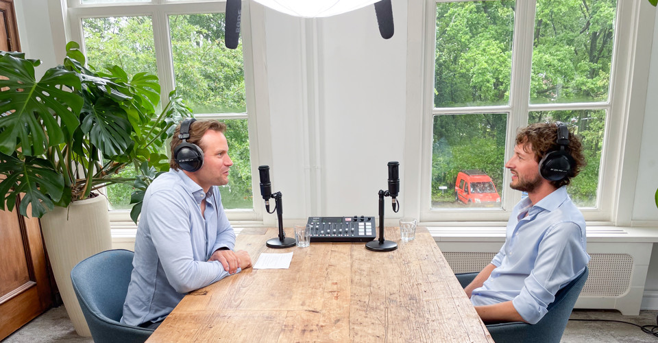 Twee mannen zitten tegenover elkaar aan een houten tafel met microfoons. Op de achtergrond zijn groene bomen te zien.