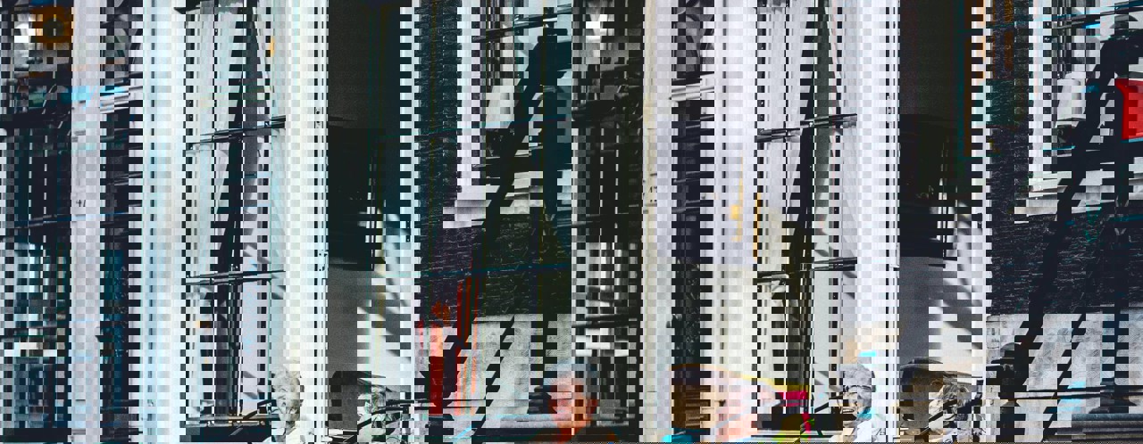 Twee personen zitten op een stoep voor een gebouw met grote ramen, met een schilderij zichtbaar op de achtergrond.