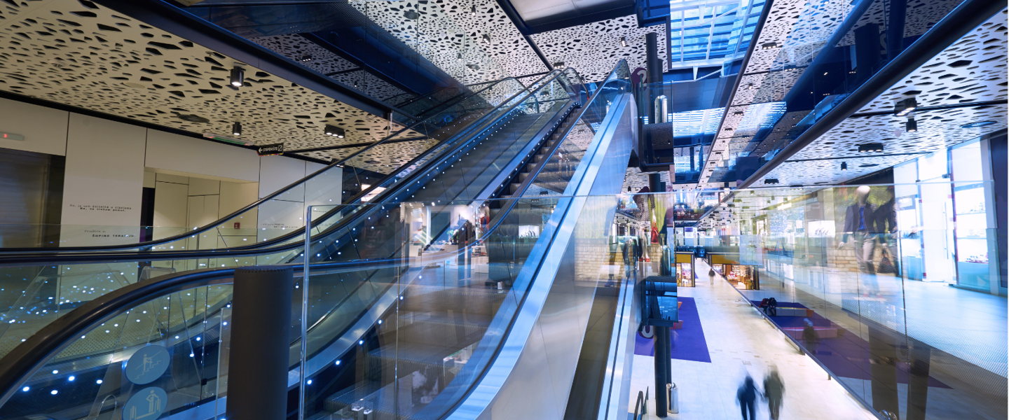 Modern shopping gallery with wide escalators, glass walls, and colorful overhead lighting, bustling with shoppers.