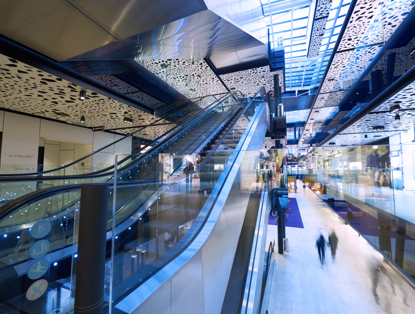 Modern shopping gallery with wide escalators, glass walls, and colorful overhead lighting, bustling with shoppers.