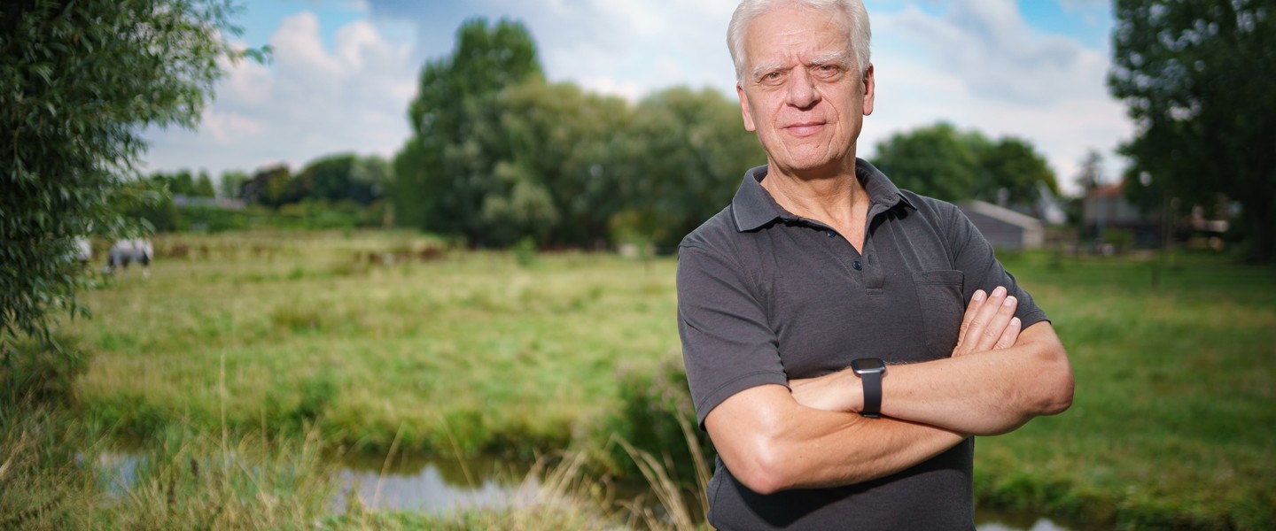 Man met gekruiste armen staat op een groene open plek naast een waterloop, met bomen en huizen op de achtergrond.