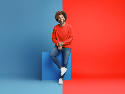 Persoon in een felrode trui zit op een blauwe kubus, met een heldere blauwe achtergrond aan de linkerkant en een heldere rode achtergrond aan de rechterkant.