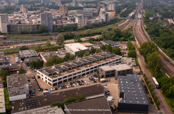 Luchtfoto van een bedrijfspark met verschillende gebouwen en parkeerplaatsen, gelegen naast een spoorlijn en in een stedelijke omgeving.
