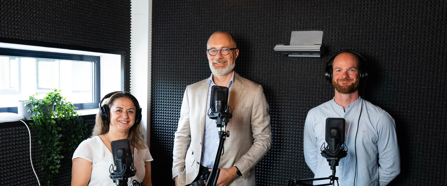 Drie presentatoren in een opnamestudio met microfoons, elk in een actieve houding, klaar om te beginnen met opnemen.