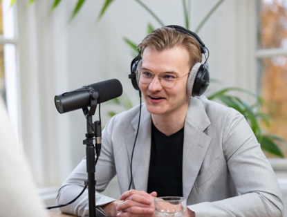 Jelle in grijze blazer en koptelefoon, zittend aan tafel met microfoon, geconcentreerd op een gesprek. Groene plant op de achtergrond.