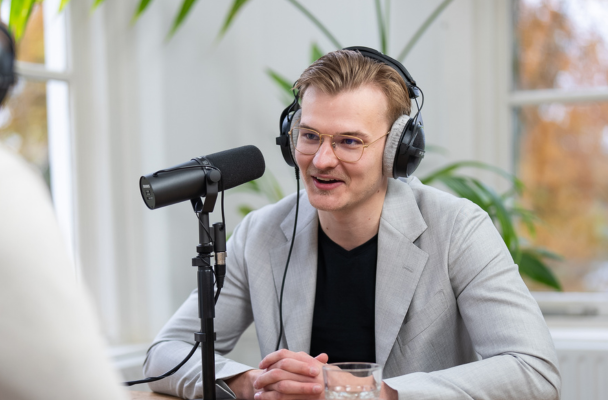 Jelle in grijze blazer en koptelefoon, zittend aan tafel met microfoon, geconcentreerd op een gesprek. Groene plant op de achtergrond.