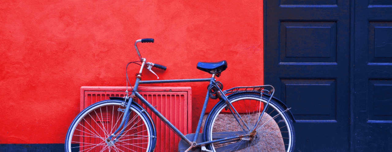 Een oude fiets leunt tegen een felrode muur naast een donkere deur, met het gebouw op de achtergrond.