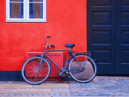 Een oude fiets leunt tegen een felrode muur naast een donkere deur, met het gebouw op de achtergrond.