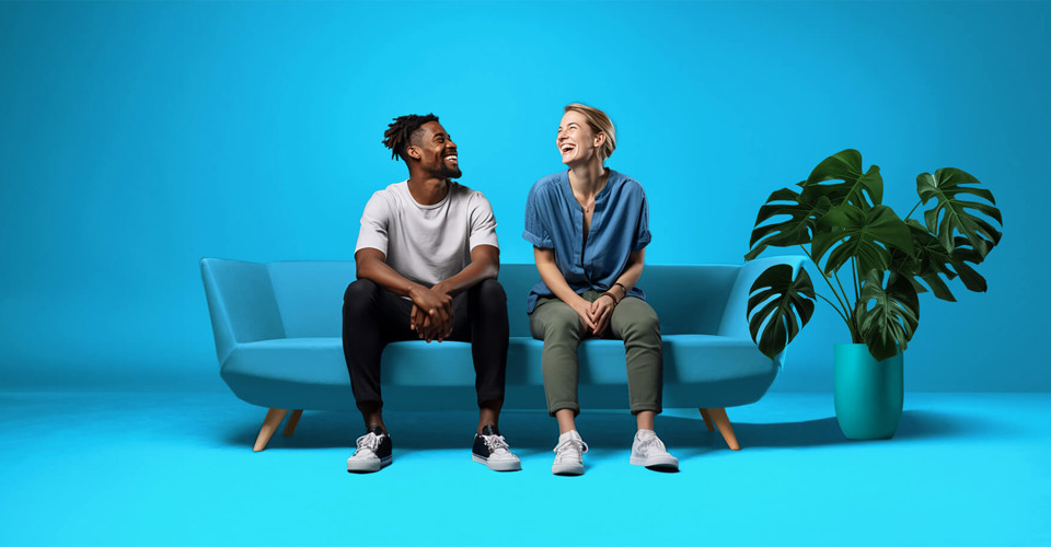 Twee mensen zitten op een lichtblauwe bank voor een felblauwe achtergrond, omringd door een groene plant.