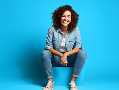 Vrouw in een spijkeroutfit zit ontspannen tegen een blauwe achtergrond.