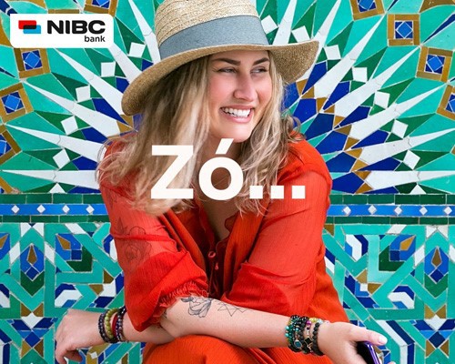 Een vrouw in een rode outfit zit voor een kleurrijke, geometrische wanddecoratie met de tekst ZÓ... in het midden.