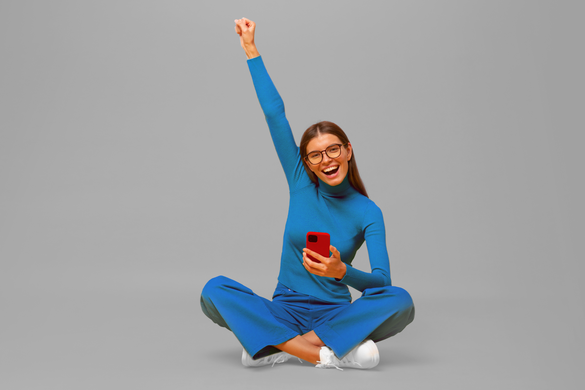 Een vrouw in een blauwe outfit zit op de grond met één arm omhoog terwijl ze een smartphone vasthoudt, stralend van enthousiasme.