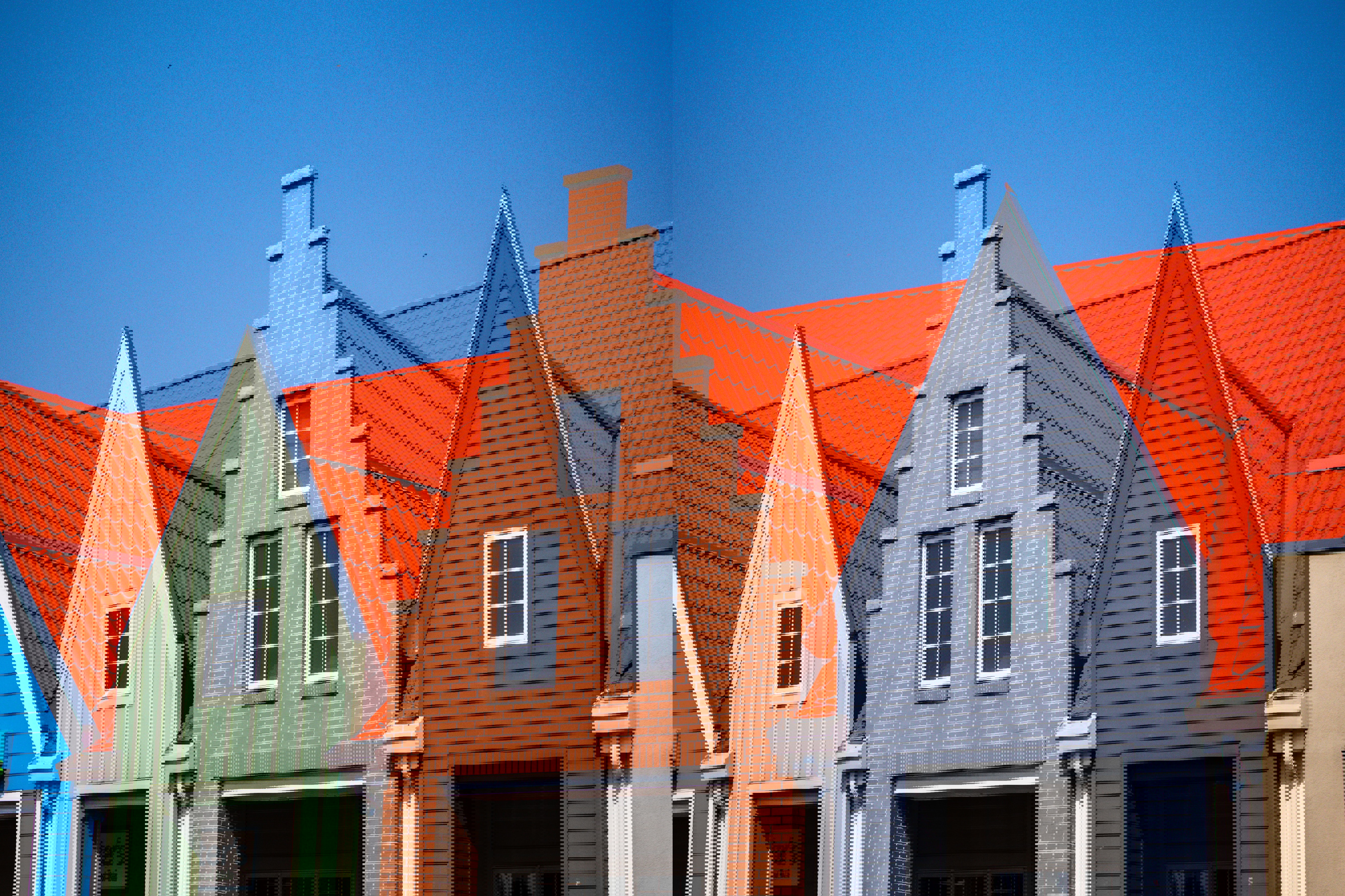 Drie naast elkaar gelegen gebouwen met verschillende gevels; het middelste gebouw heeft een bakstenen muur en een hoog puntdak, omringd door gebouwen met groene en grijze gevels en oranje daken.