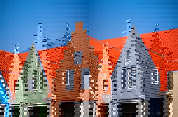 Drie naast elkaar gelegen gebouwen met verschillende gevels; het middelste gebouw heeft een bakstenen muur en een hoog puntdak, omringd door gebouwen met groene en grijze gevels en oranje daken.