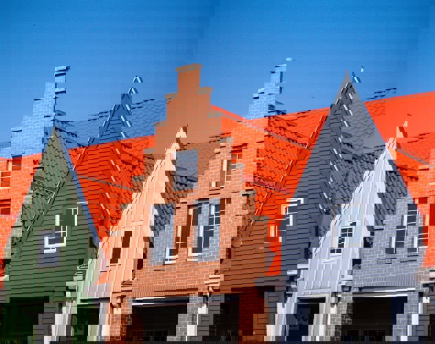 Drie naast elkaar gelegen gebouwen met verschillende gevels; het middelste gebouw heeft een bakstenen muur en een hoog puntdak, omringd door gebouwen met groene en grijze gevels en oranje daken.