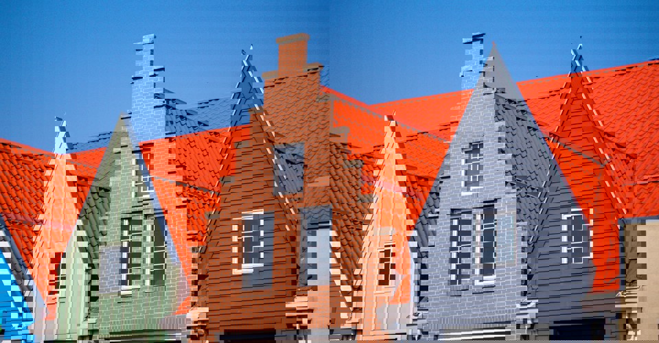 Drie naast elkaar gelegen gebouwen met verschillende gevels; het middelste gebouw heeft een bakstenen muur en een hoog puntdak, omringd door gebouwen met groene en grijze gevels en oranje daken.