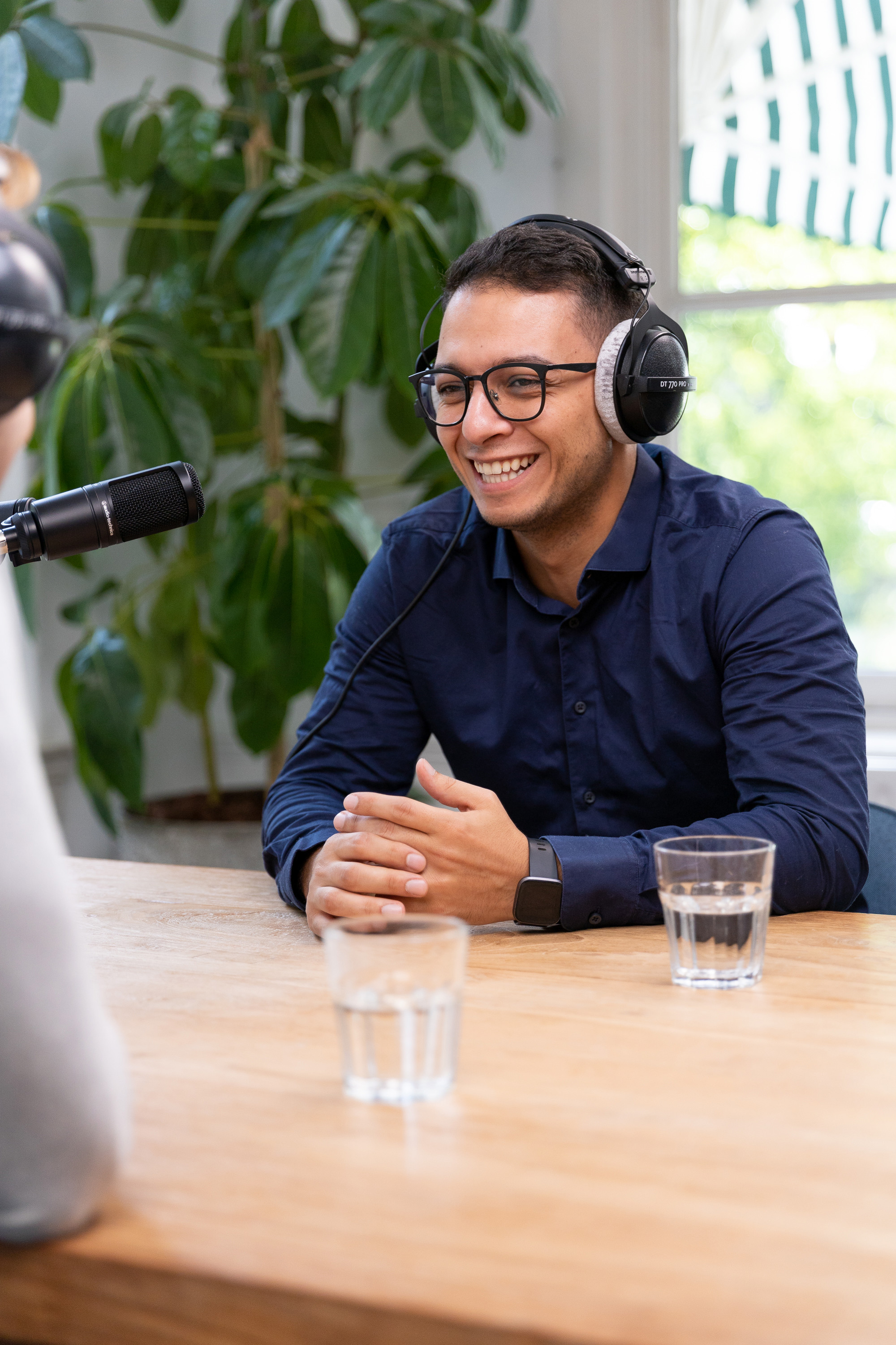 Twee mensen zitten aan een houten tafel, met een microfoon, hoorapparatuur en glazen water, terwijl ze een gesprek of interview voeren.