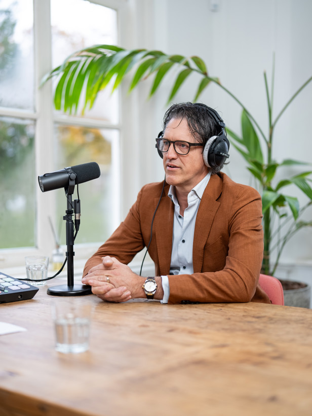 Wijnand in een bruine blazer zit aan een tafel met een koptelefoon op, voor hem staat een microfoon en op de achtergrond is een plant te zien.