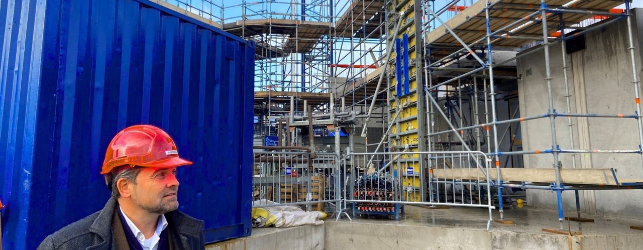 Een bouwplaats met steiger en containers, waar een persoon in een bouwhelm staat.