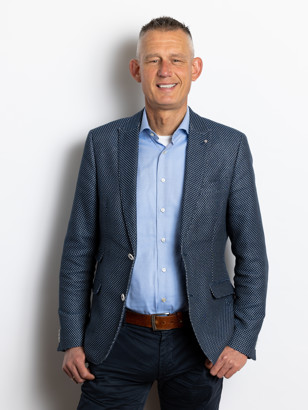 Stefan De Wit in een gestreepte blazer en een lichtblauw overhemd staat met zijn handen in zijn zakken voor een neutrale achtergrond.