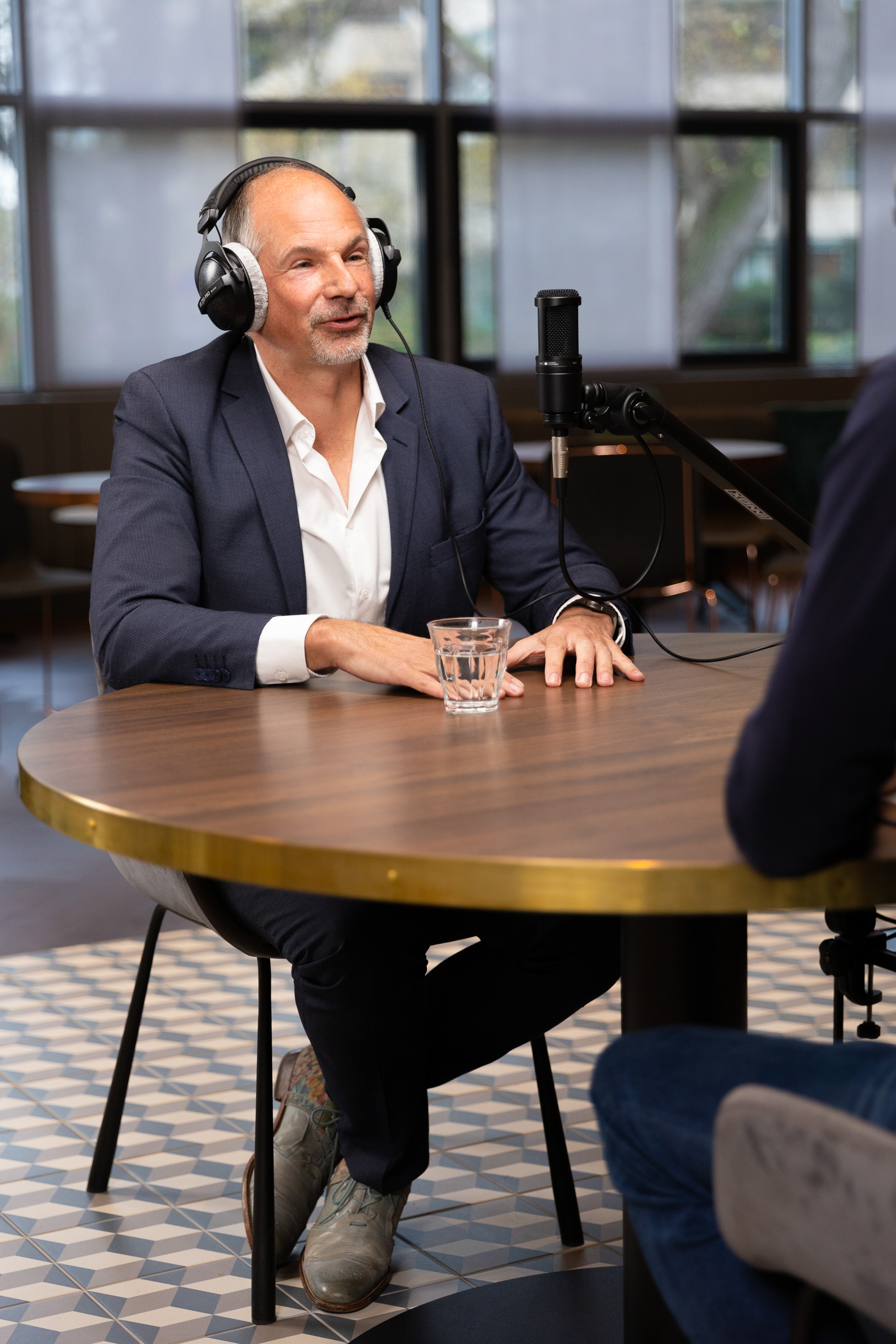 Man in pak met een headset, zittend aan een tafel met een glas water, in gesprek met iemand buiten beeld.