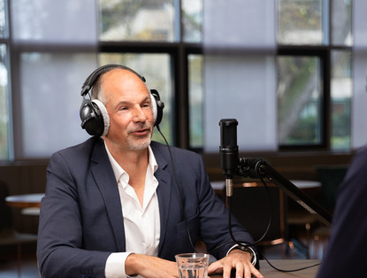 Man in pak met een headset, zittend aan een tafel met een glas water, in gesprek met iemand buiten beeld.