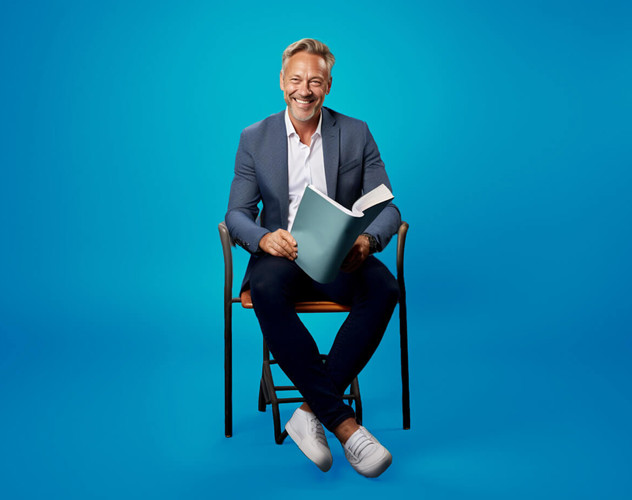 Man in een blazer zit op een stoel met een boek in zijn handen, voor een heldere blauwe achtergrond.