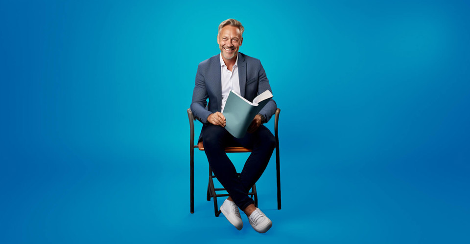 Man in een blazer zit op een stoel met een boek in zijn handen, voor een heldere blauwe achtergrond.