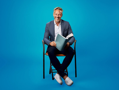 Man in een blazer zit op een stoel met een boek in zijn handen, voor een heldere blauwe achtergrond.