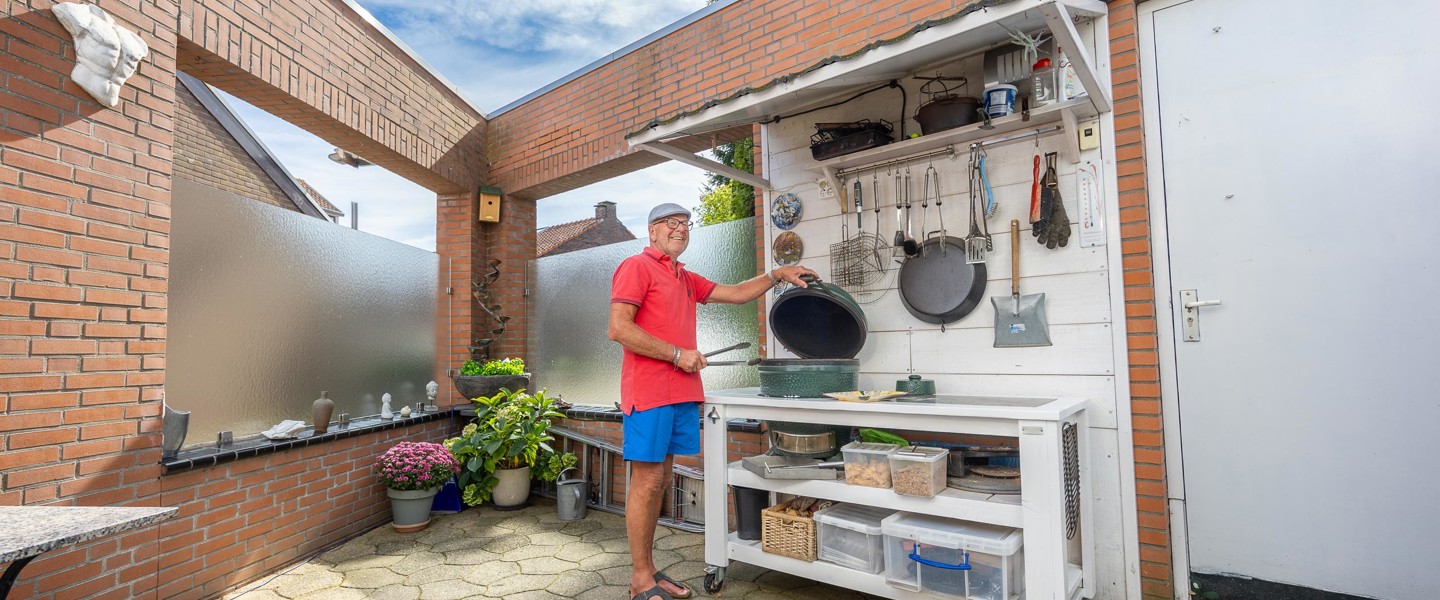 Man in een rood shirt en blauwe shorts staat bij een buitenkeuken, omringd door kookgerei en kruidenplanten.