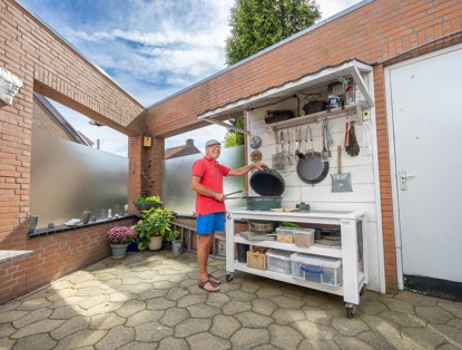 Man in een rood shirt en blauwe shorts staat bij een buitenkeuken, omringd door kookgerei en kruidenplanten.
