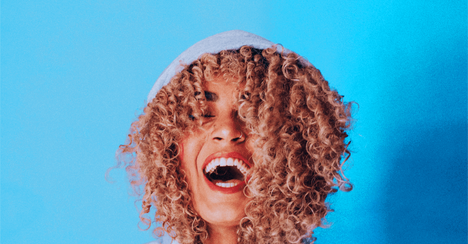 Persoon met krullend haar in een grijze hoodie, staand voor een blauwe achtergrond.