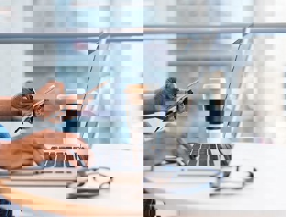 Een persoon met zwart gelakte nagels typt op een laptop terwijl hij/zij een smartphone vasthoudt, met een koffiebeker naast zich.