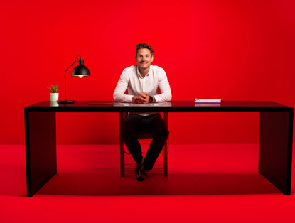 Een man in een wit shirt zit achter een zwart bureau met een tafelblad, voor een felrode achtergrond.