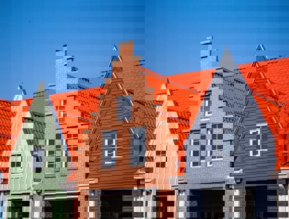 Drie naast elkaar gelegen gebouwen met verschillende gevels; het middelste gebouw heeft een bakstenen muur en een hoog puntdak, omringd door gebouwen met groene en grijze gevels en oranje daken.